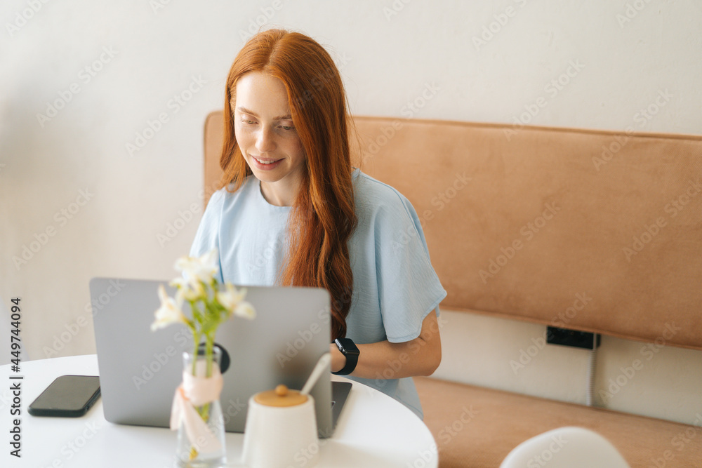 Medium shot of positive attractive young woman student working on project using laptop sitting at desk in cozy cafe, looking on screen. Pretty redhead Caucasian lady remote working or studying. Medium shot of positive attractive young woman student working on project using laptop sitting at desk in cozy cafe, looking on screen. Pretty redhead Caucasian lady remote working or studying.