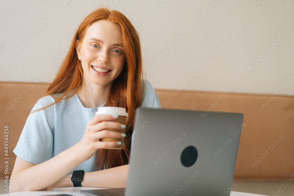 Close-up face of happy young beautiful woman holding cup with hot coffee sitting at desk with laptop at cozy cafe, looking at camera. Pretty redhead Caucasian lady remote working or studying. Close-up face of happy young beautiful woman holding cup with hot coffee sitting at desk with laptop at cozy cafe, looking at camera. Pretty redhead Caucasian lady remote working or studying.