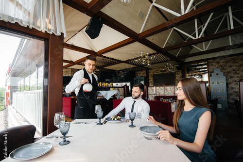 A young waiter in a stylish apron serves a table with a beautiful couple in a refined restaurant. Customer service.