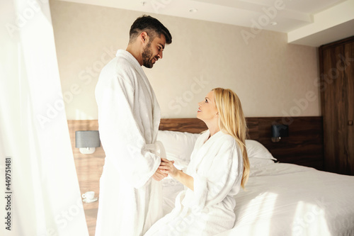 Love attraction and respect. Waking up in a warm hotel with natural light. A man stands above the bed and gently holds the girl's hands. She sits on the bed, they both have white bathrobes on. Loving