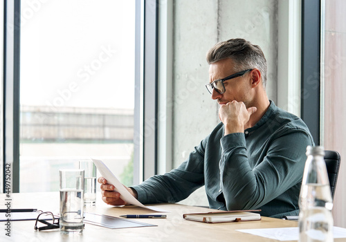 Focused serious mature businessman professional lawyer executive holding paper checking document analyzing financial report, male legal expert banker reading contract sitting at office table. photo