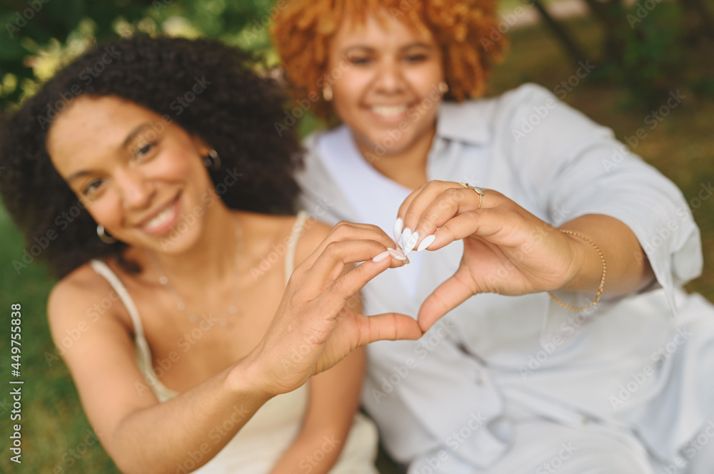 © Алина Троева - Young beautiful happy lesbian African American couple sitting on green grass showing love sign outside at nature summer park. LGBT community concept. Female friends smiling enjoying moments together. © Алина Троева - Young beautiful happy lesbian African American couple sitting on green grass showing love sign outside at nature summer park. LGBT community concept. Female friends smiling enjoying moments together.