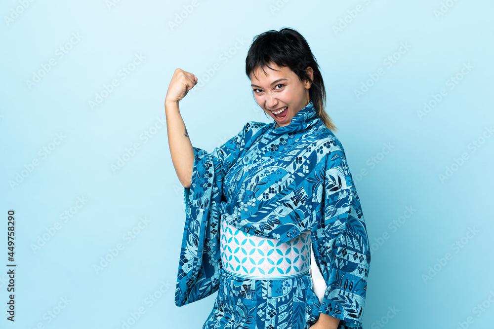 Young woman wearing kimono over isolated blue background celebrating a victory Young woman wearing kimono over isolated blue background celebrating a victory