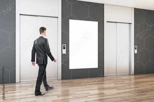 Businessman walking in modern office lobby interior with steel elevators, empty white poster, wooden flooring and tile wall. Mock up.