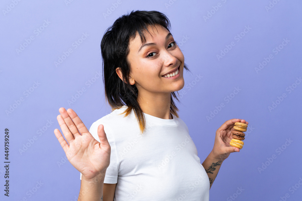 Young Uruguayan woman over isolated purple background holding colorful French macarons and saluting Young Uruguayan woman over isolated purple background holding colorful French macarons and saluting