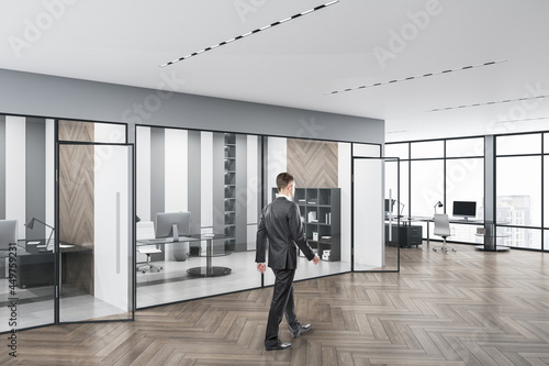 Business man walking in modern concrete glass office interior with daylight, equipment and wooden flooring. Workplace design concept.