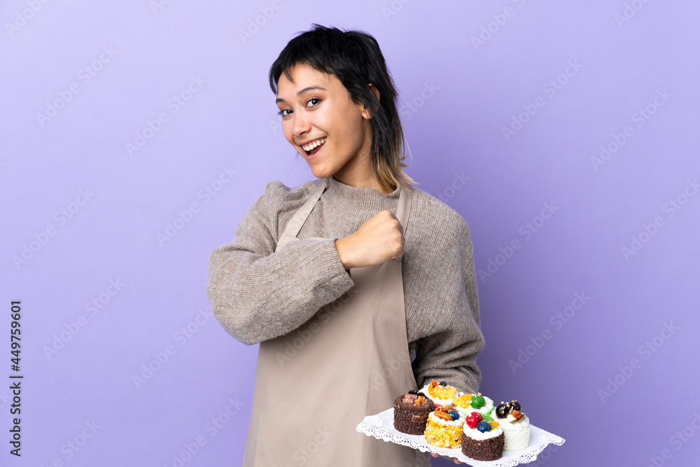 Young Uruguayan woman holding lots of different mini cakes over isolated purple background celebrating a victory Young Uruguayan woman holding lots of different mini cakes over isolated purple background celebrating a victory