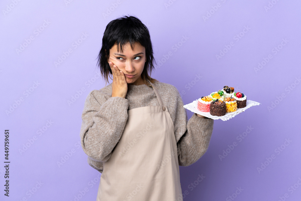Young Uruguayan woman holding lots of different mini cakes over isolated purple background unhappy and frustrated Young Uruguayan woman holding lots of different mini cakes over isolated purple background unhappy and frustrated