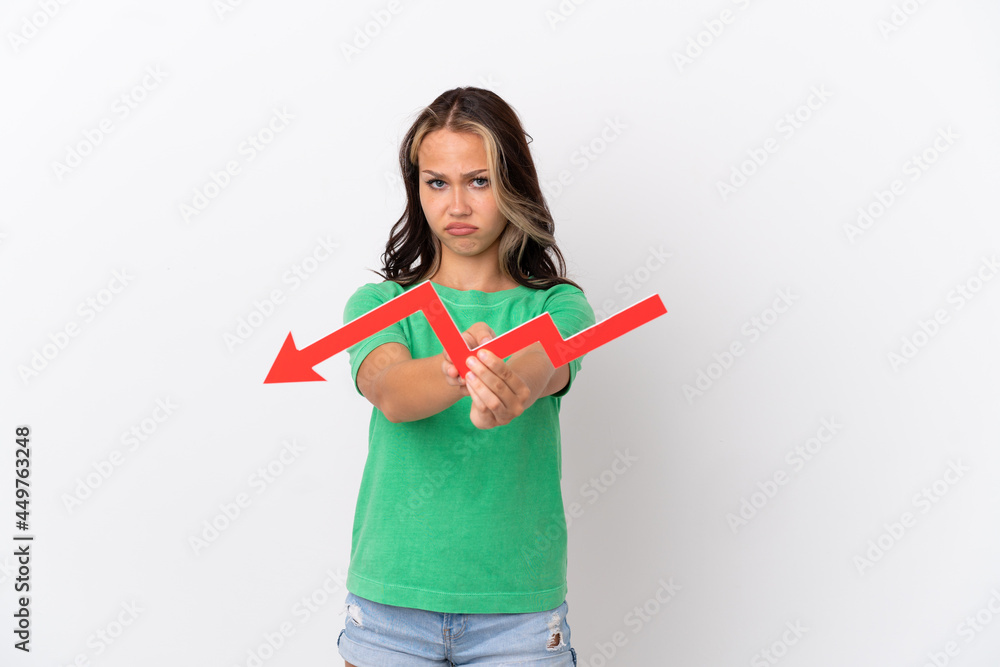 Teenager Russian girl isolated on blue background holding a downward arrow and with sad expression Teenager Russian girl isolated on blue background holding a downward arrow and with sad expression