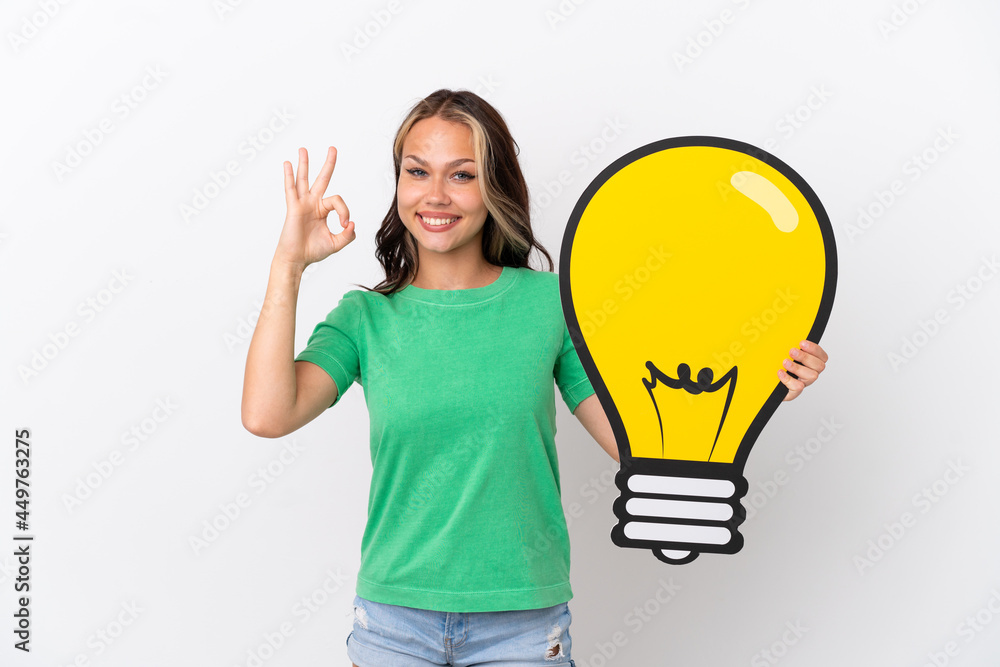 Teenager Russian girl isolated on blue background holding a bulb icon and doing OK sign Teenager Russian girl isolated on blue background holding a bulb icon and doing OK sign