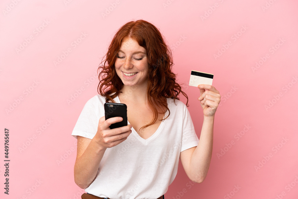 Teenager reddish woman isolated on pink background buying with the mobile with a credit card Teenager reddish woman isolated on pink background buying with the mobile with a credit card