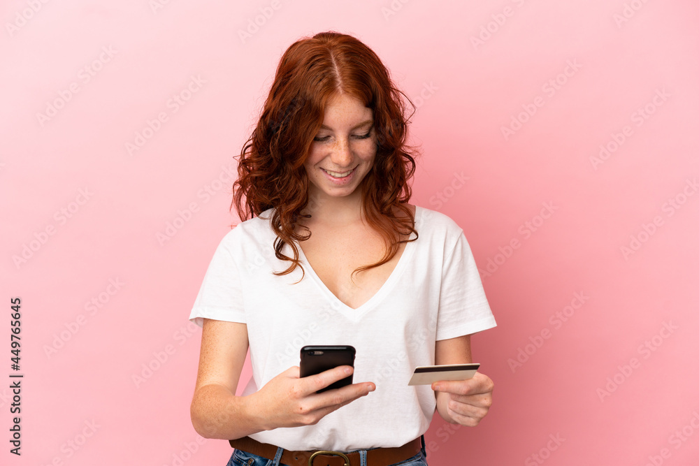 Naklejka premium Teenager reddish woman isolated on pink background buying with the mobile with a credit card