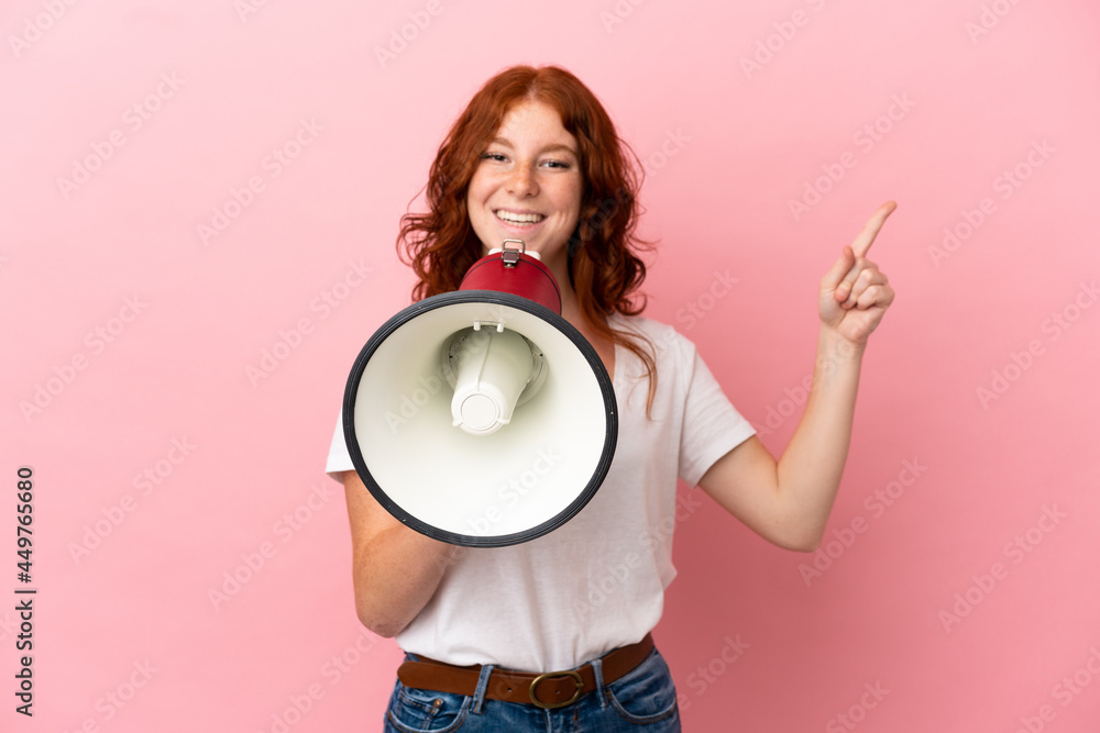 Teenager reddish woman isolated on pink background shouting through a megaphone and pointing side Teenager reddish woman isolated on pink background shouting through a megaphone and pointing side