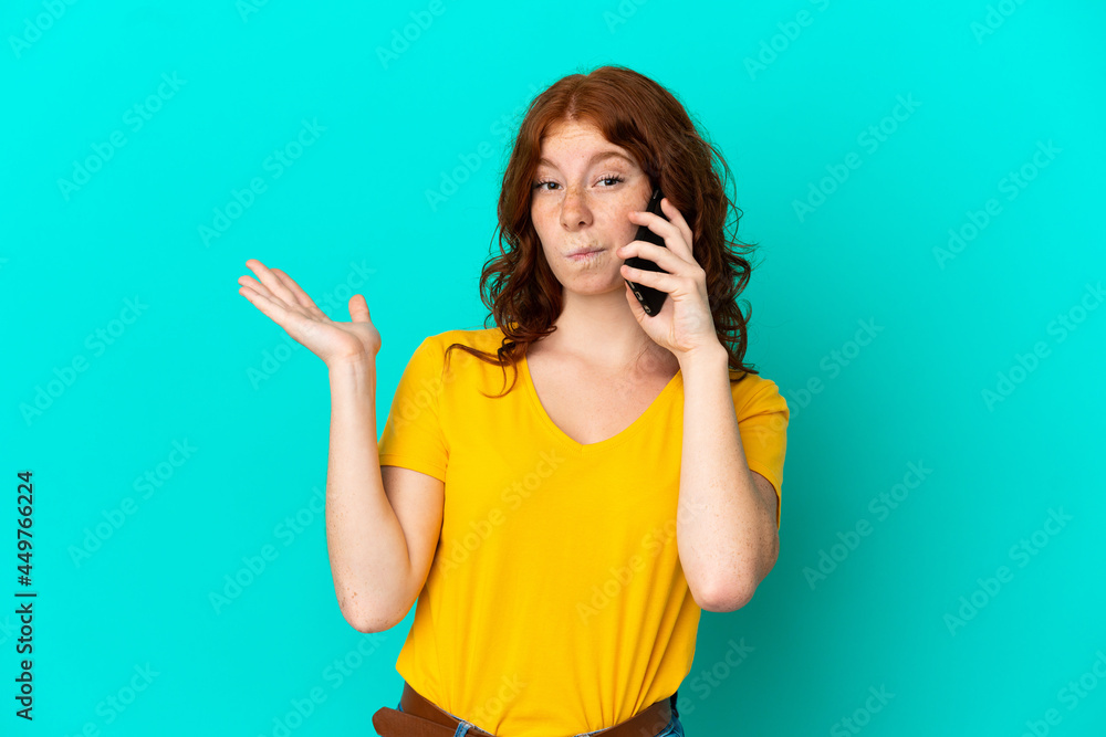 Teenager reddish woman using mobile phone isolated on blue background having doubts while raising hands Teenager reddish woman using mobile phone isolated on blue background having doubts while raising hands