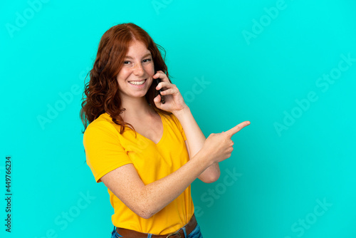 Teenager reddish woman using mobile phone isolated on blue background pointing finger to the side