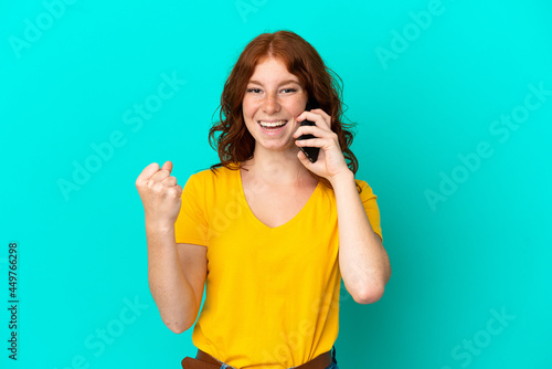 Teenager reddish woman using mobile phone isolated on blue background celebrating a victory in winner position