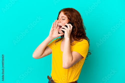 Teenager reddish woman using mobile phone isolated on blue background shouting with mouth wide open to the side