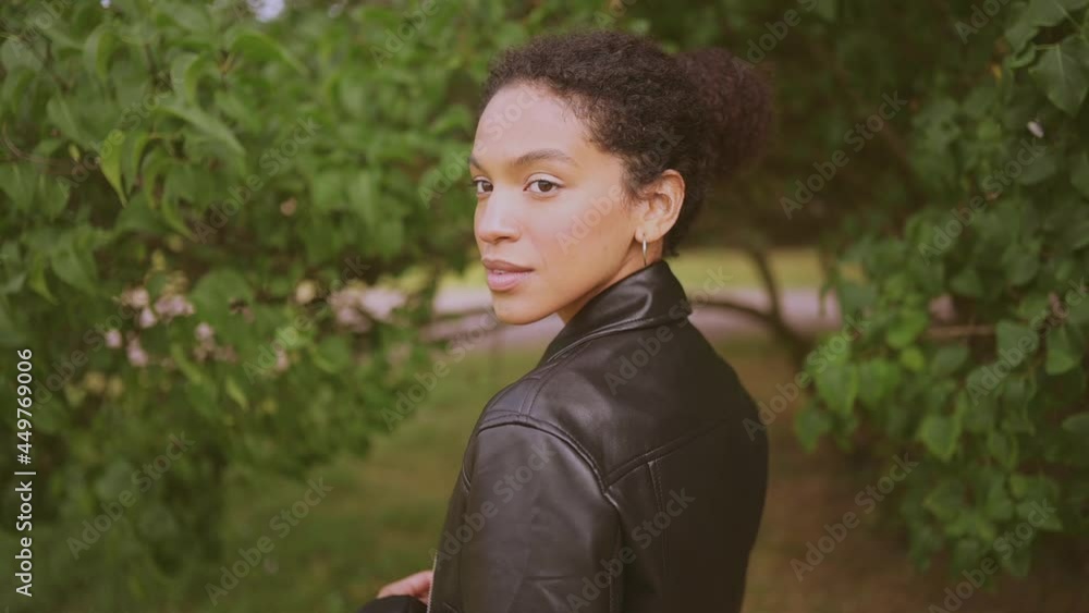 Fashion close up slow motion portrait of sensual attractive young naturally beautiful African American woman with afro hair in black leather jacket posing in nature parkland in green foliage Fashion close up slow motion portrait of sensual attractive young naturally beautiful African American woman with afro hair in black leather jacket posing in nature parkland in green foliage
