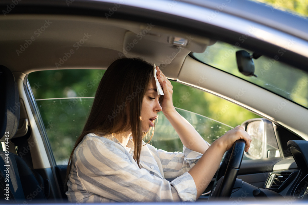 Girl driver being hot during heat wave in car, suffering from hot weather, has problem with a non-working air conditioner, wipes sweat from her face with tissue. Summer, heat concept. Girl driver being hot during heat wave in car, suffering from hot weather, has problem with a non-working air conditioner, wipes sweat from her face with tissue. Summer, heat concept.