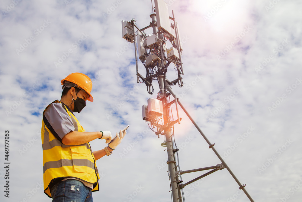 Asian male worker engineer wearing a helmet and safety goggles uses a smartphone to field work near a telecommunication tower controlling cellular electrical installations. - obrazy, fototapety, plakaty 