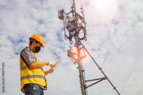 Asian male worker engineer wearing a helmet and safety goggles uses a smartphone to field work near a telecommunication tower controlling cellular electrical installations.