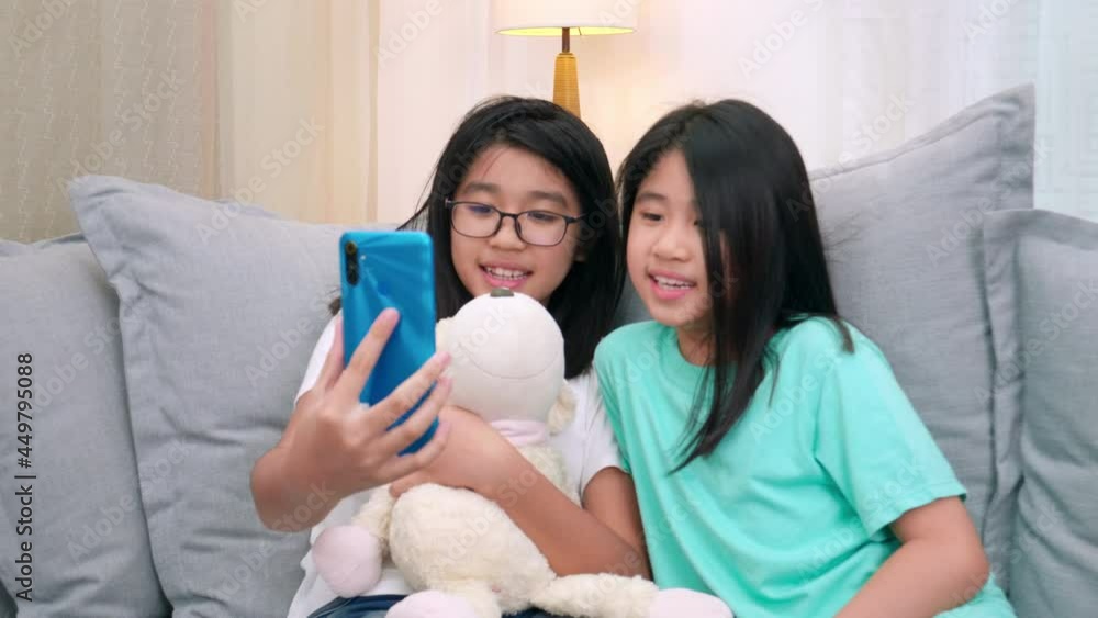 Happy siblings children sitting on sofa in living room speak with parent on mobile phone together, smiling elder girl showing cute little sister video call by smartphone speak with family.. Lifestyle Happy siblings children sitting on sofa in living room speak with parent on mobile phone together, smiling elder girl showing cute little sister video call by smartphone speak with family.. Lifestyle