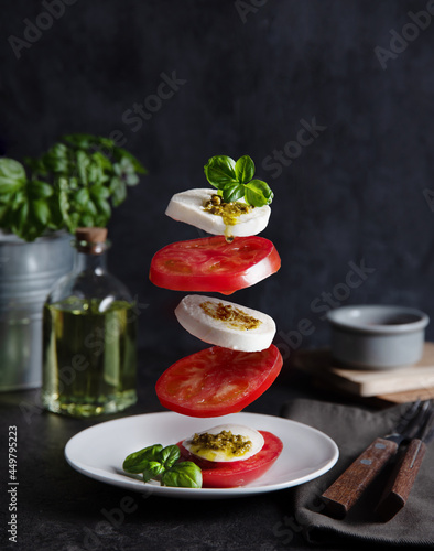 Concept levitation food. Italian caprese salad with tomatoes, mozzarella cheese, basil and pesto sauce fly over the plate on a dark background. Front view photo