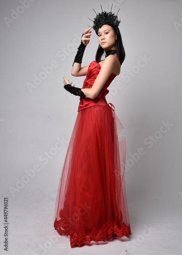 Full length portrait of beautiful young asian woman wearing red corset and ornate gothic queen crown. Graceful standing posing isolated on studio background.