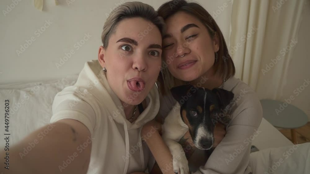 Lgbt couple kissing and taking selfie with dog on couch in home room indoors spbd. Lgbt couple kissing and taking selfie with dog on couch in home room indoors spbd.