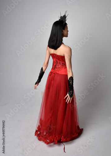 Full length portrait of beautiful young asian woman wearing red corset and ornate gothic queen crown. Graceful standing posing isolated on studio background.