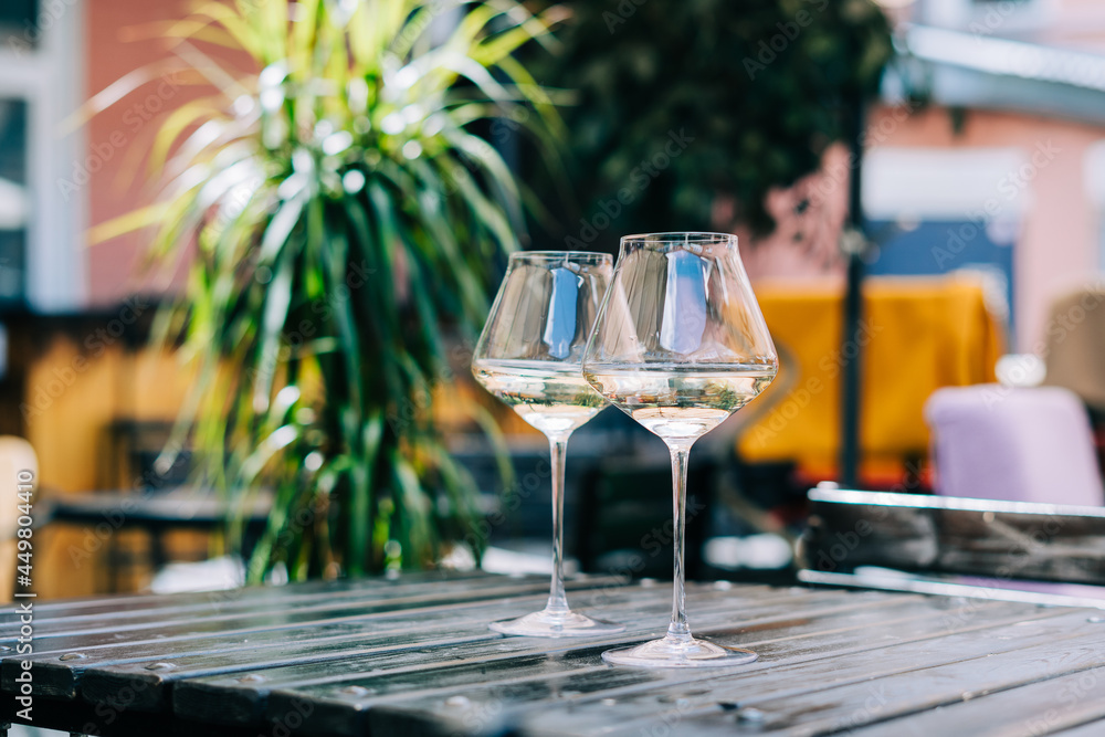 A wooden table in a restaurant with white wine. Wine glasses served for a party in a bar or a restaurant on terrace at a sunny day. Blurred background with plants. Still life scene A wooden table in a restaurant with white wine. Wine glasses served for a party in a bar or a restaurant on terrace at a sunny day. Blurred background with plants. Still life scene