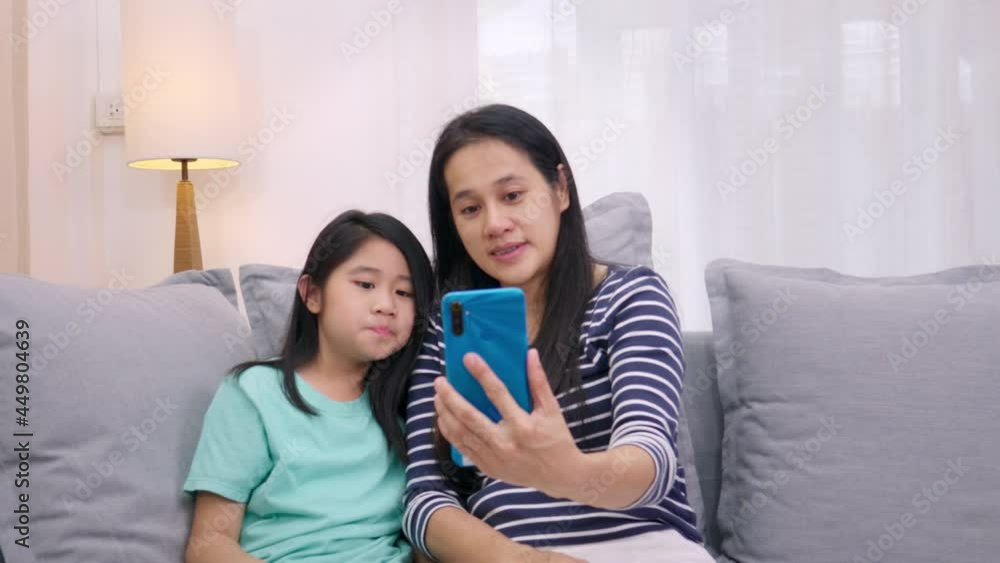 Happy Asian family mother and daughter sitting on sofa talking video conference by smartphone together in living room at home. mother and cute little girl video call by smartphone.Technology lifestyle Happy Asian family mother and daughter sitting on sofa talking video conference by smartphone together in living room at home. mother and cute little girl video call by smartphone.Technology lifestyle