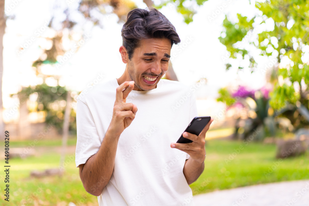 Fototapeta premium Young caucasian man in a park using mobile phone with fingers crossing