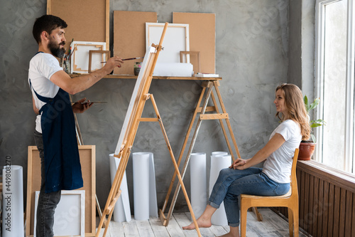 male artist dressed in a blue dirty apron stands in the studio barefoot and paints a woman's portrait. Girl model in casual clothes posing for him.