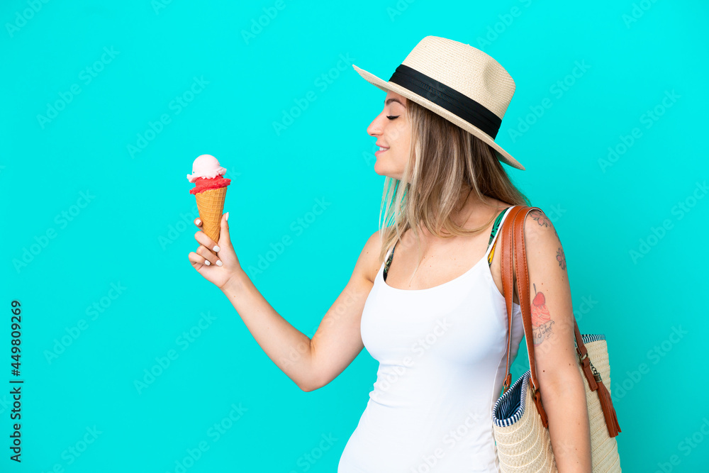 Young Romanian woman holding ice cream and beach bag isolated on blue background with happy expression Young Romanian woman holding ice cream and beach bag isolated on blue background with happy expression