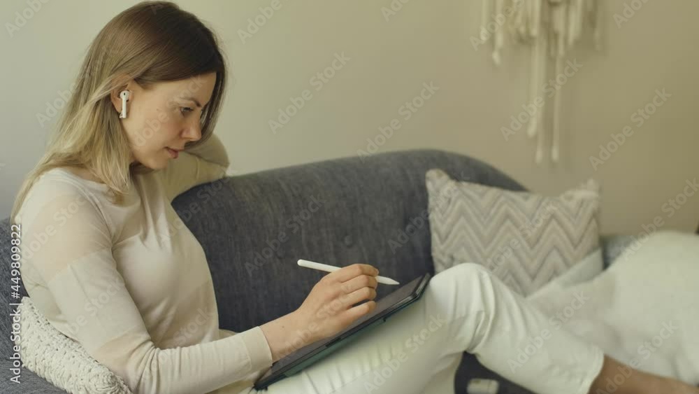 Woman with tablet using pencil tapping and painting at home on sofa. Lying on the couch and listen to music. Online remote working on tablet. Out of office due covid. Girl coloring sheets on tablet. Woman with tablet using pencil tapping and painting at home on sofa. Lying on the couch and listen to music. Online remote working on tablet. Out of office due covid. Girl coloring sheets on tablet.