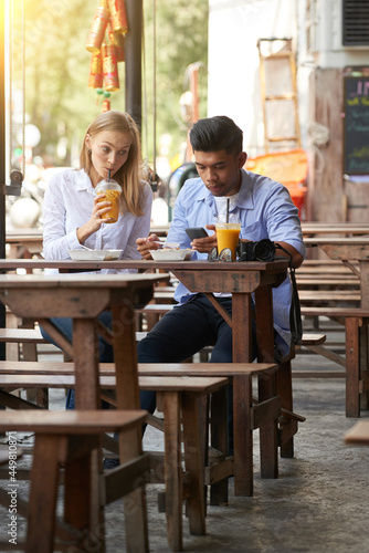 Fototapeta Naklejka Na Ścianę i Meble -  Young woman drinking refreshing cocktail at outdoor cafe and looking at boyfriend texting friends or using app on smartphone