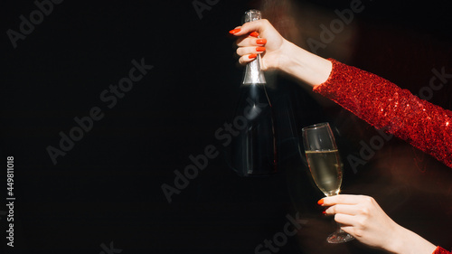 Christmas spirit. Party joy. Festive celebration. Advertising background. Female hands red spangle dress holding glass proposing bottle of champagne long exposure blur light on black copy space.