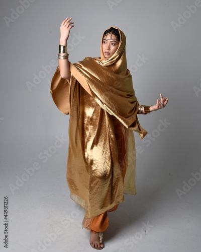Full length portrait of pretty young asian woman wearing golden Arabian robes like a genie, standing pose holding flowing fabric, isolated on studio background.