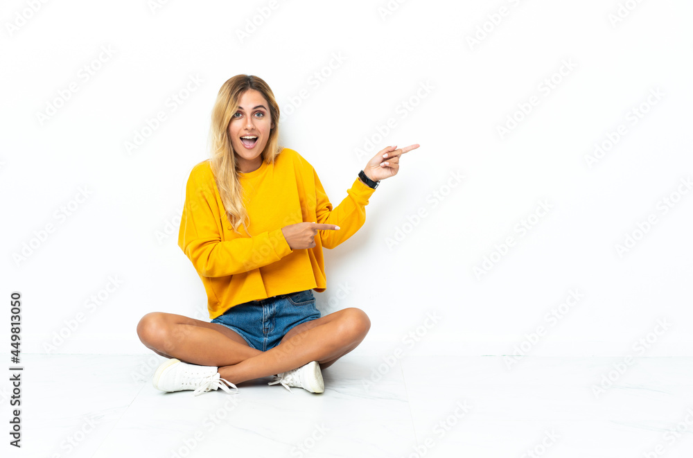 Young blonde Uruguayan woman sitting on the floor isolated on white background surprised and pointing side Young blonde Uruguayan woman sitting on the floor isolated on white background surprised and pointing side