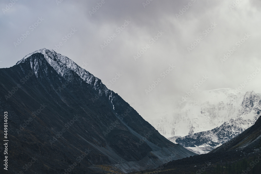 Dark atmospheric landscape with high black mountain top and snow-covered mountain wall in low clouds. Dramatic foggy mountain landscape with peaked top and snowy mountains in fog in overcast weather.