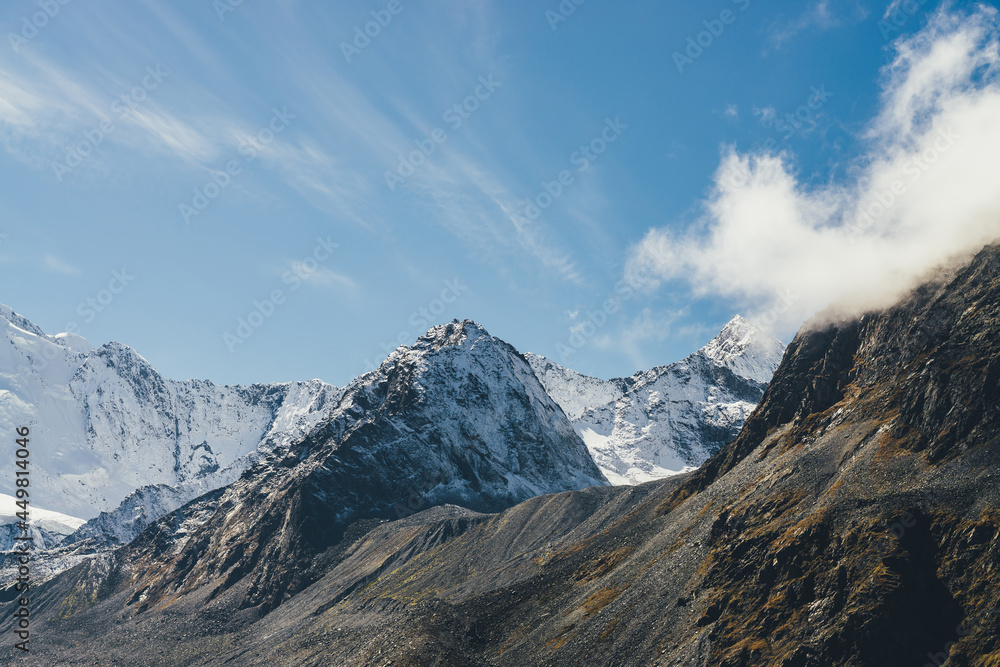 Atmospheric alpine landscape with high snowy mountain with peaked top under cirrus clouds in sky. Big snow covered mountain in sunshine. Low cloud on black rocks and white-snow pointy peak in sunlight