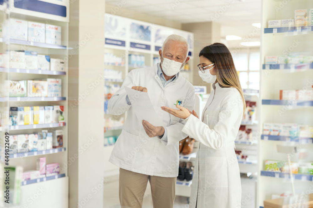 Education in pharmacy and corona virus. A mature pharmacist explains the documentation in the pharmacy to a young pharmacist in professional training. They wear uniforms and face masks