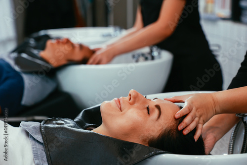 Serious Asian female hairdresser wearing uniform and spectacles shampooing hair of young woman customer in modern salon using organic shampoo and conditioner