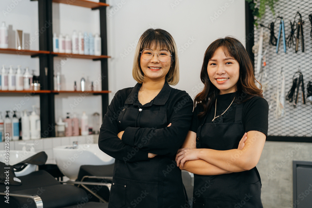 Portrait of team of happy successful young Asian hairstylist standing wearing apron and spectacles with folded hands smiling while looking at camera Portrait of team of happy successful young Asian hairstylist standing wearing apron and spectacles with folded hands smiling while looking at camera