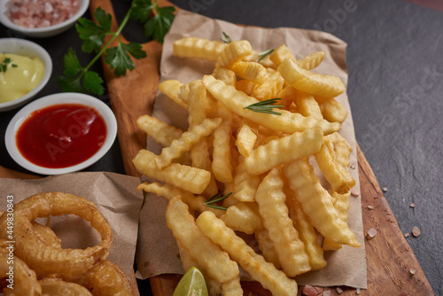 Homemade baked potato fries with mayonnaise, Tomato sauce and rosemary on wooden board. fast food products : onion rings, french fries on cutting board, on black stone background, unhealthy food.
