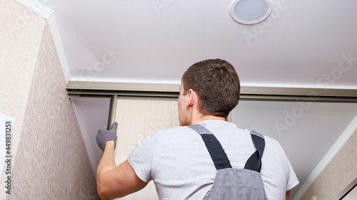 Employee furniture assembler in white t-shirt and jumpsuit checks wooden sliding door of modern wardrobe with mirror close back low angle shot