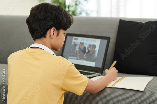 Fototapeta Naklejka Na Ścianę i Meble -  Serious concentrated teenage boy attending online class and writing in copybook