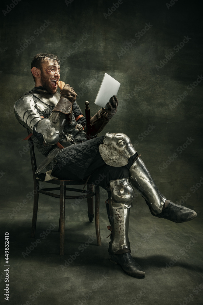 One brutal bearded man, medeival warrior or knight with digital tablet sitting on chair over dark background. - obrazy, fototapety, plakaty 