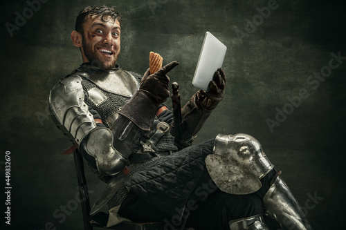 One brutal bearded man, medeival warrior or knight with digital tablet sitting on chair over dark background.